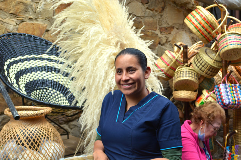 Una mujer artesana sonríe mientras se encuentra en un mercado rodeada de canastos tejidos y productos tradicionales. Su trabajo refleja la herencia cultural y la importancia de las labores artesanales que hacen parte del registro vivo de las comunidades en el censo