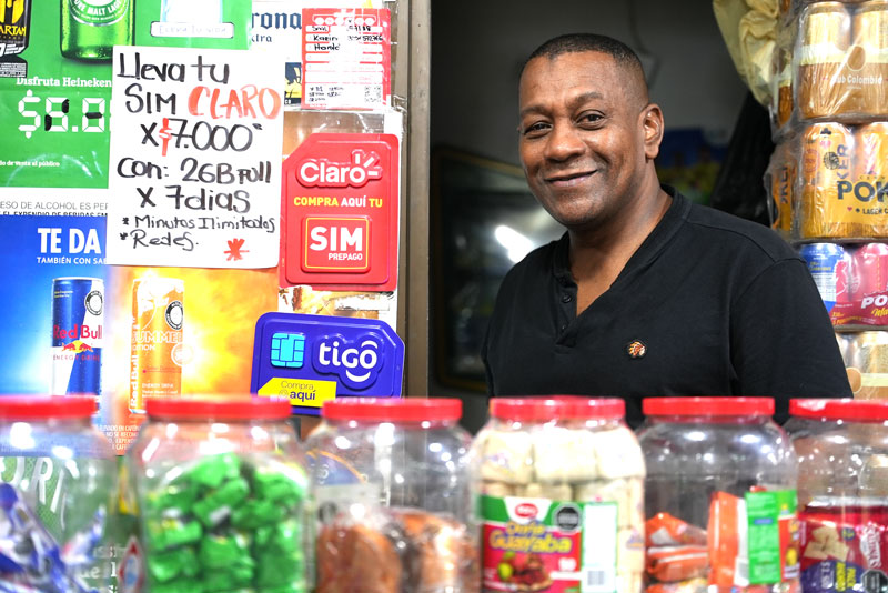 
                            En su pequeña tienda de barrio, un hombre sonriente atiende a los clientes entre frascos de dulces y productos básicos. A su alrededor, carteles de recargas y tarjetas SIM evidencian el dinamismo del comercio local