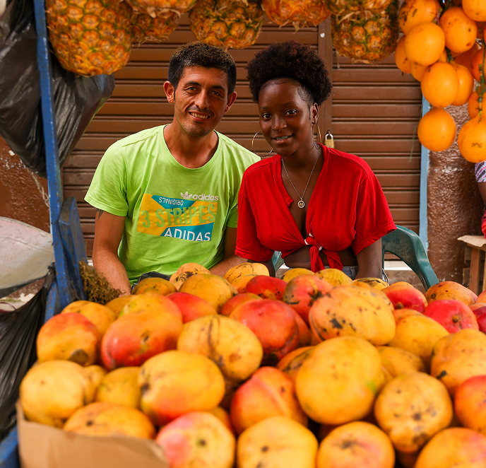 Un hombre y una mujer sonriendo detrás de un puesto de mercado lleno de mangos y otras frutas colgando al fondo