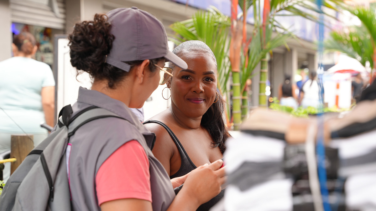 Una censista del DANE que lleva una gorra y un chaleco con el logo de la entidad hablando con otra mujer que sonríe, en lo que parece ser un entorno de mercado o calle